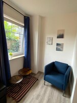 A room with a blue armchair, a small round table, a window with curtains, and two wall-mounted pictures.
