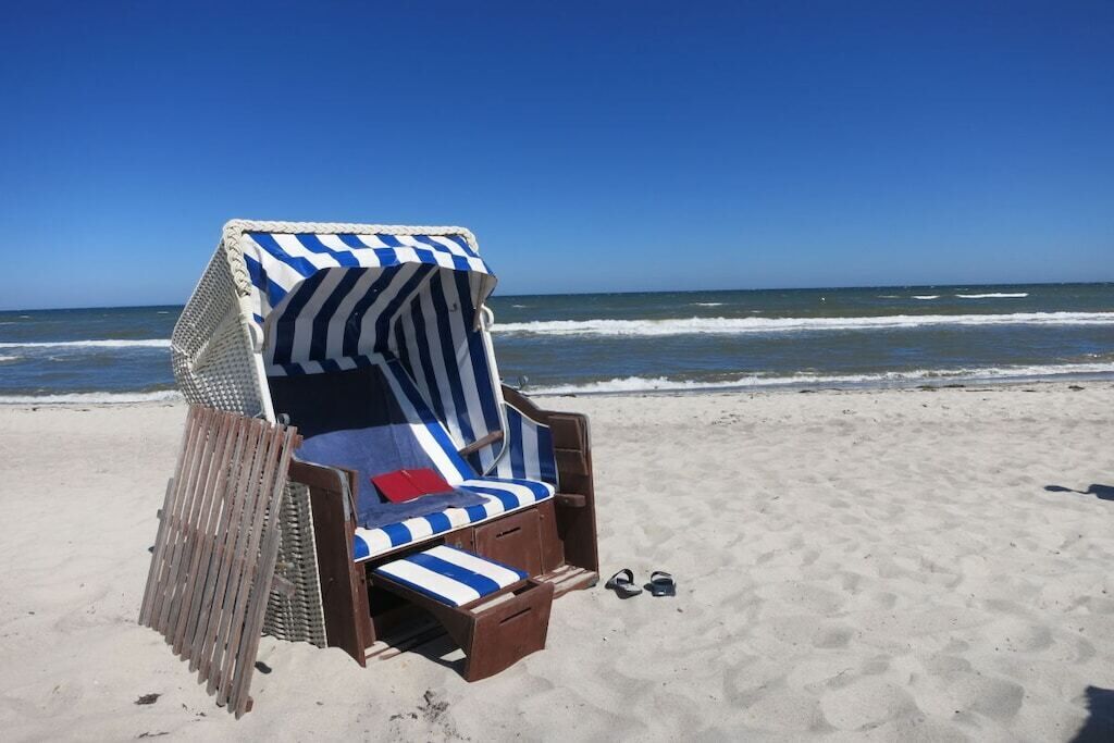 Une chaise de plage avec un revêtement à rayures, une paire de chaussures et une clôture en bois sur une plage de sable, avec l’océan en arrière-plan.
