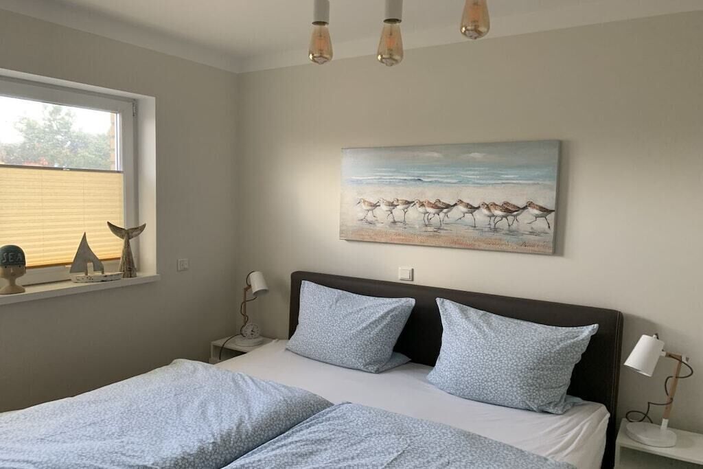 A bedroom with a bed, two bedside tables, a window with blinds, a painting of birds on the wall, and a ceiling light fixture.
