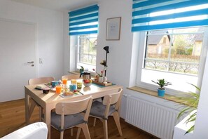 Een eetgedeelte met een tafel voor twee personen, een radiator onder de ramen en een plant in een pot op de vensterbank.