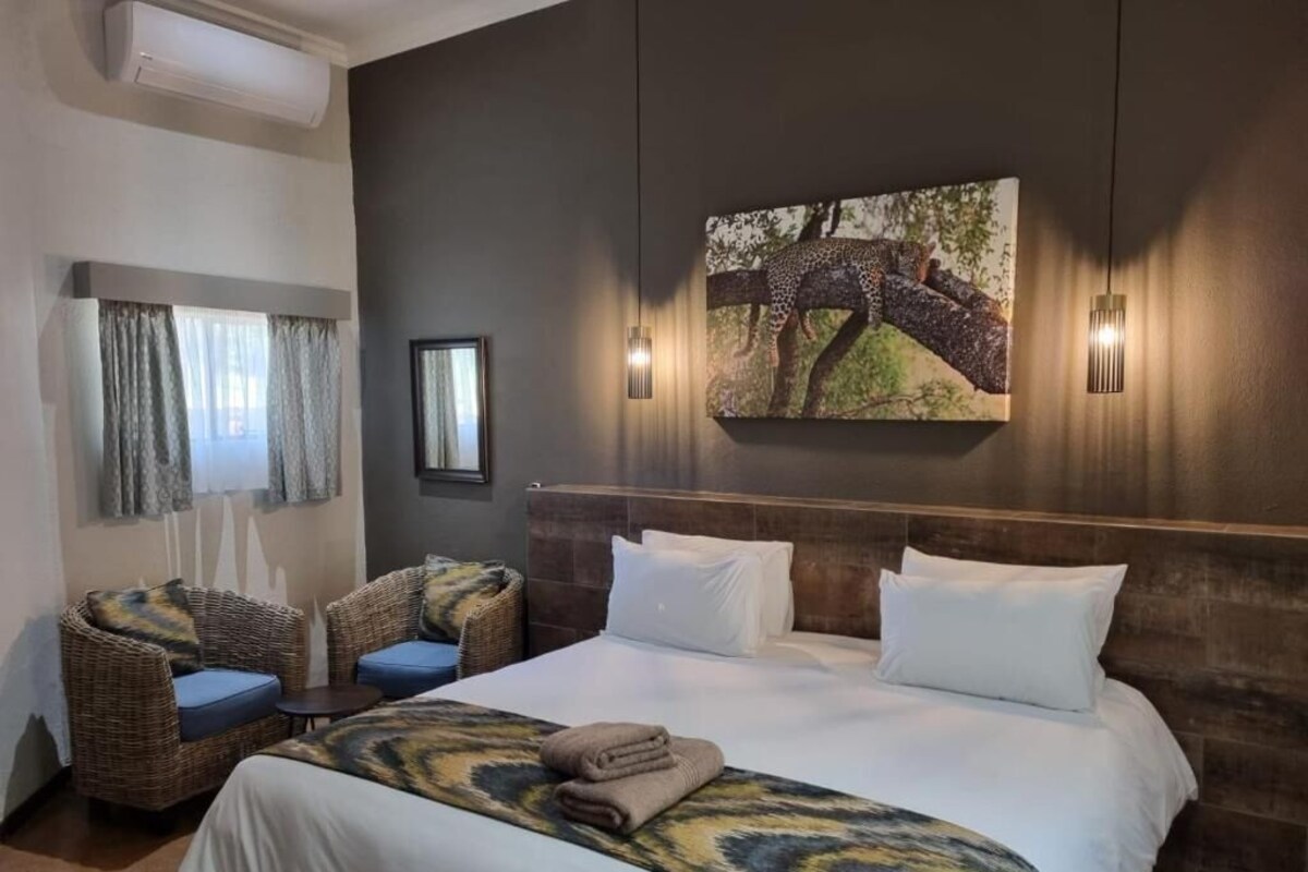 A hotel room with a large bed, two wicker chairs with blue cushions, a painting of a leopard on a tree, and a wall-mounted light fixture.