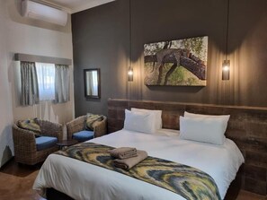 A hotel room with a large bed, two wicker chairs with blue cushions, a painting of a leopard on a tree, and a wall-mounted light fixture. - Kruger Whispering Waters (Hazyview)