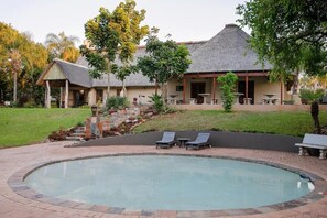 A pool area with lounge chairs, a thatched-roof building, and a landscaped garden. - Kruger Whispering Waters (Hazyview)