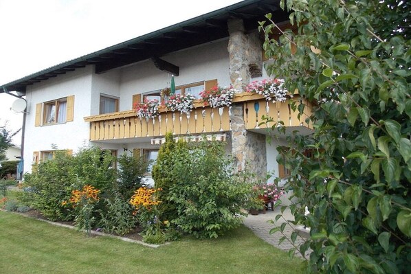 Ein zweigeschossiges Gebäude mit einem Balkon, der mit Blumen geschmückt ist, einer Steinsäule und einem gepflegten Garten mit verschiedenen Pflanzen und Blumen.