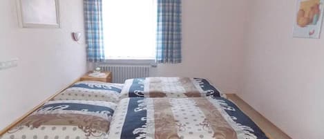 A single bed with a patterned bedspread and pillows, a bedside table, a window with curtains, and a radiator.