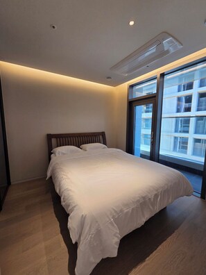 A modern bedroom with a large bed, a window with a view of buildings, and recessed ceiling lighting. - A brand-new, luxury high-end officetel in Seocho-dong (Seoul)