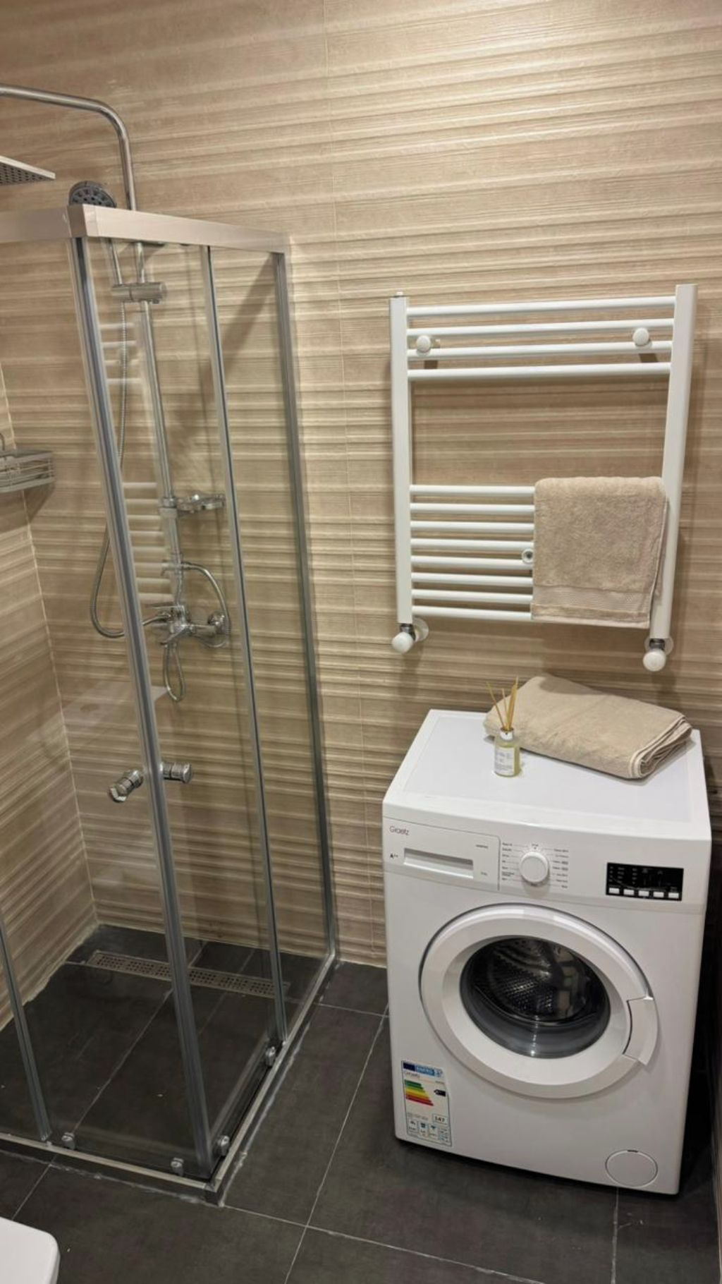 Casa de banho com cabine de duche em vidro, suporte de toalhas branco e máquina de lavar frontal.