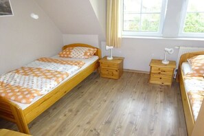 A small, single-room accommodation with a wooden floor, two beds with matching bedding, a small wooden nightstand with a lamp, and a window with curtains. - Holiday apartment with 2 bedrooms (Elbinsel Krautsand)