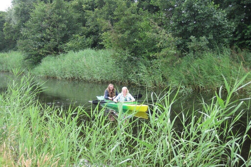 Two people in a green kayak on a calm river surrounded by tall grass and dense trees.