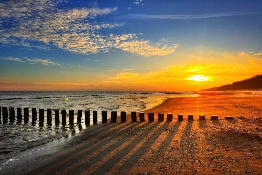 Une plage avec une jetée en bois qui s’avance dans l’eau, un ciel dégagé et le soleil qui se couche à l’horizon.