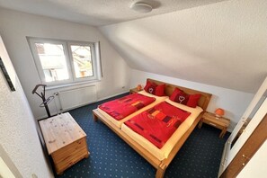 A bedroom with a sloped ceiling, a bed with red bedding, a wooden nightstand, and a window with a view of buildings. - Holiday apartment with 1 bedroom (Ückeritz)