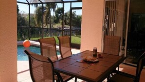 Uma área de pátio com mesa e cadeiras, um espaço envidraçado com vista para uma piscina e vegetação, e uma porta de correr.