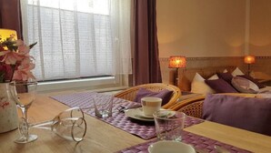 A room with a dining table set for two, a wicker chair, a bed with pillows, and a window with curtains. - "BALTIC SAND" with balcony (Sassnitz)