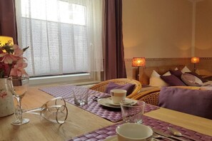Een kamer met een eettafel gedekt voor twee personen, een rieten stoel, een bed met kussens en een raam met gordijnen.