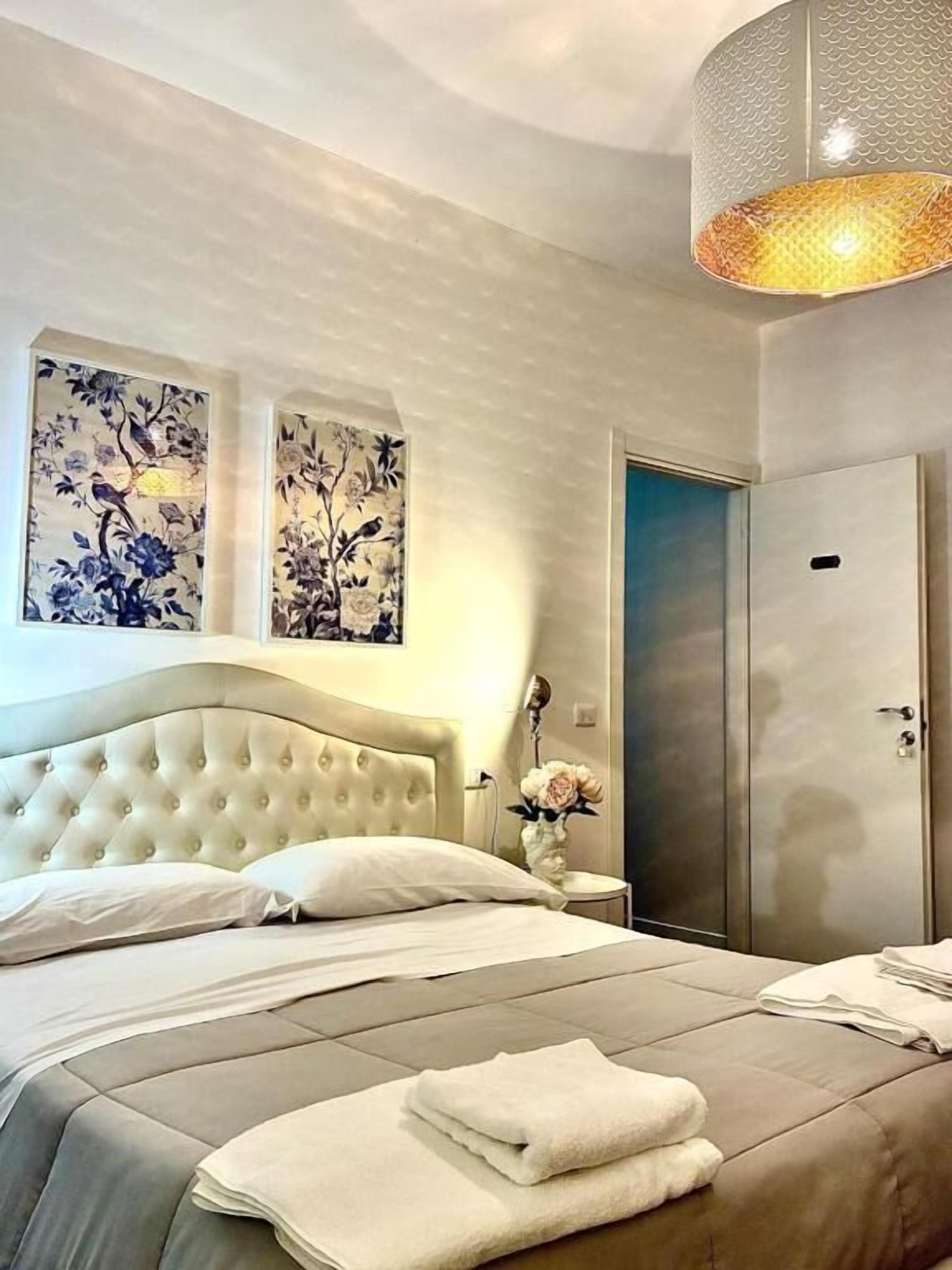 A bedroom with a tufted headboard, a bed with white linens, a bedside table with a vase, and a wardrobe.