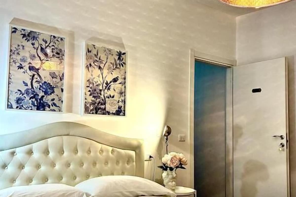 Une chambre à coucher avec une tête de lit capitonnée, un lit avec du linge de lit blanc, une table de chevet avec un vase et une armoire.