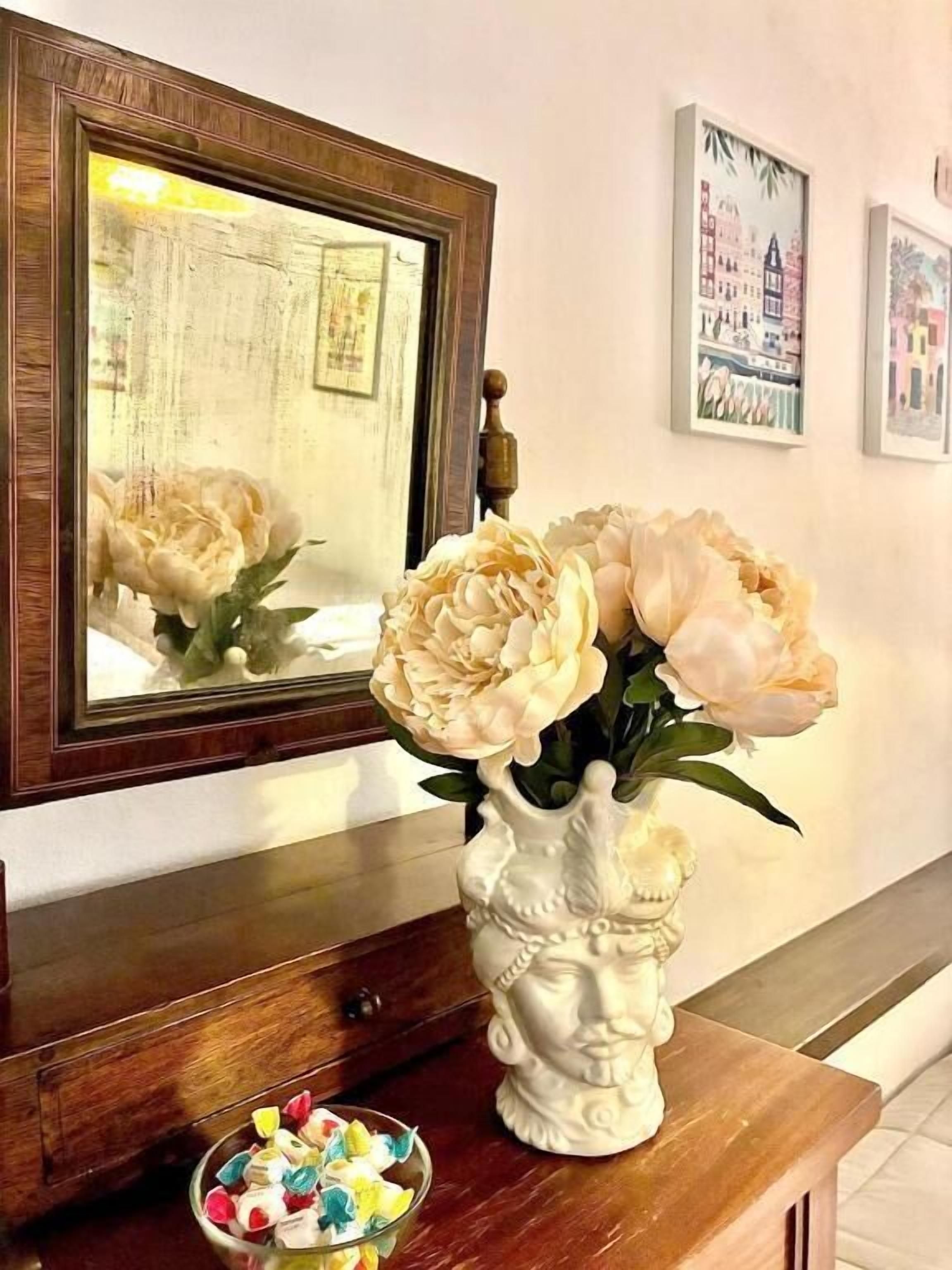 Un miroir reflétant un vase de fleurs, un petit bol rempli de bonbons colorés et des photos encadrées accrochées au mur.