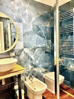 Une salle de bain avec un mur en marbre, un miroir rond, un lavabo, un bidet et des toilettes.