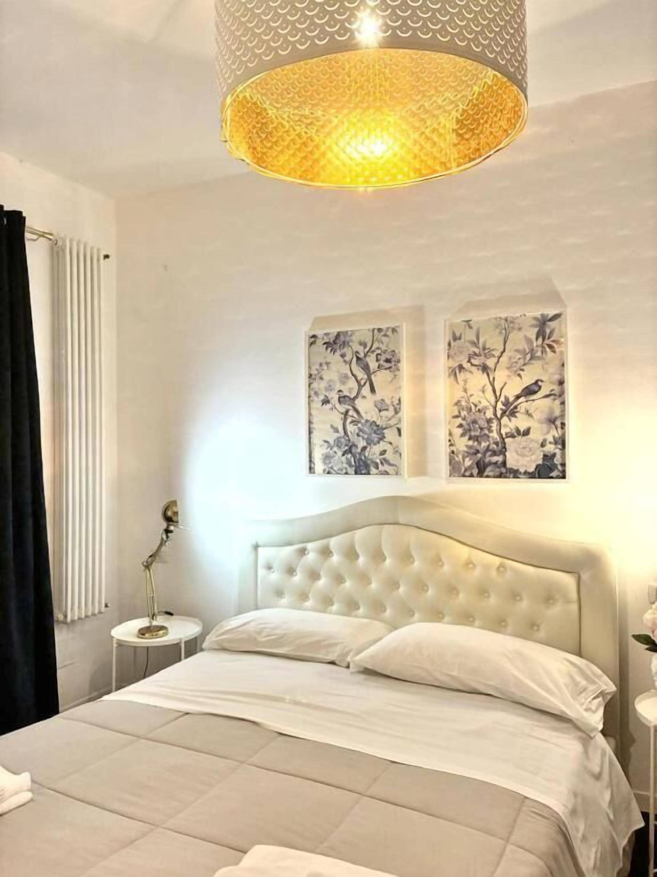Un dormitorio con cabecera capitonada, cama con ropa blanca, una mesita redonda con lámpara y dos obras de arte colgadas en la pared.