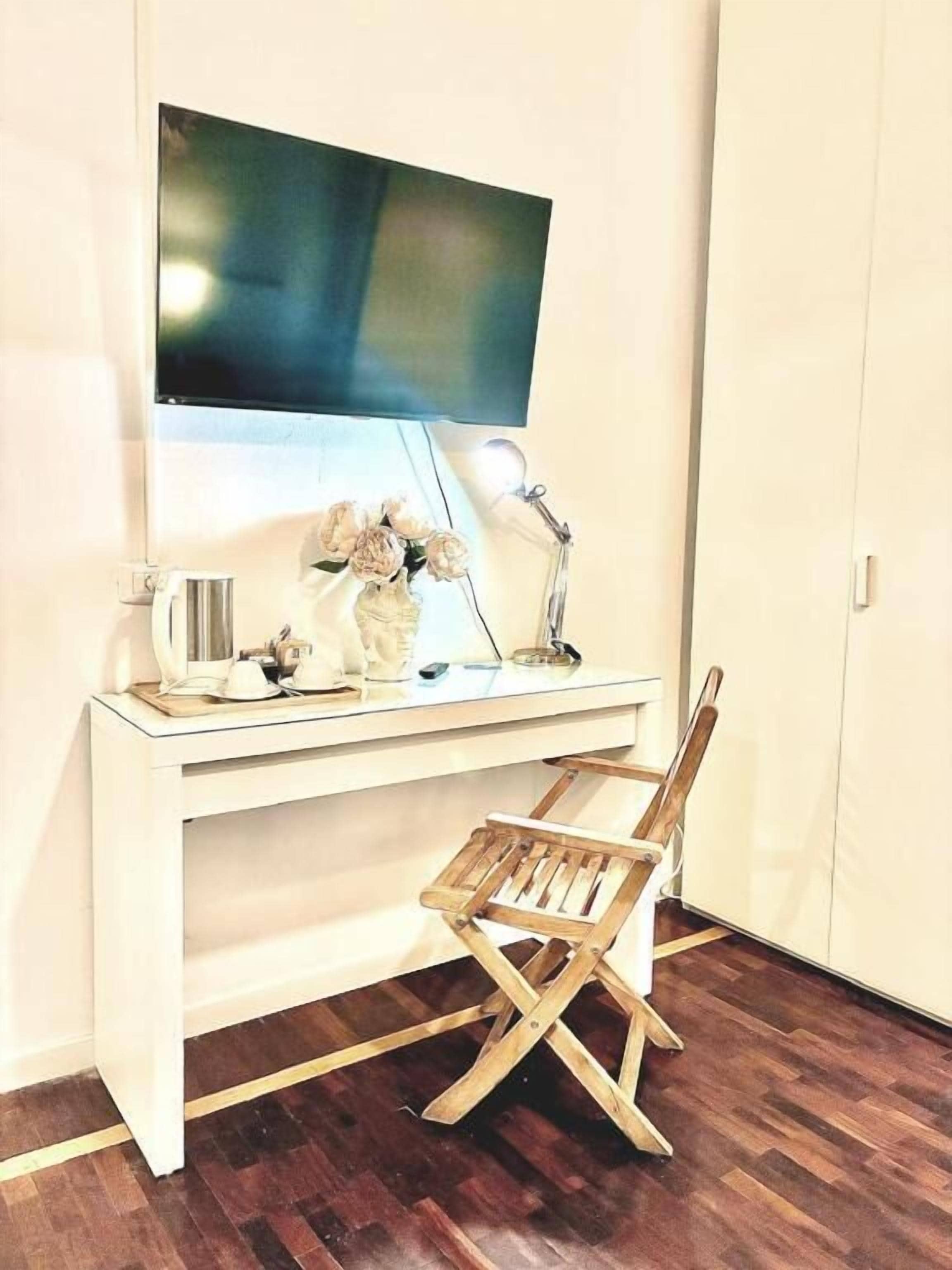Una habitación con un escritorio blanco, un televisor montado en la pared, una lámpara de escritorio, una pequeña silla y suelo de madera.