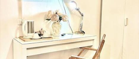 A room with a white desk, a television mounted on the wall, a desk lamp, a small chair, and a wooden floor.