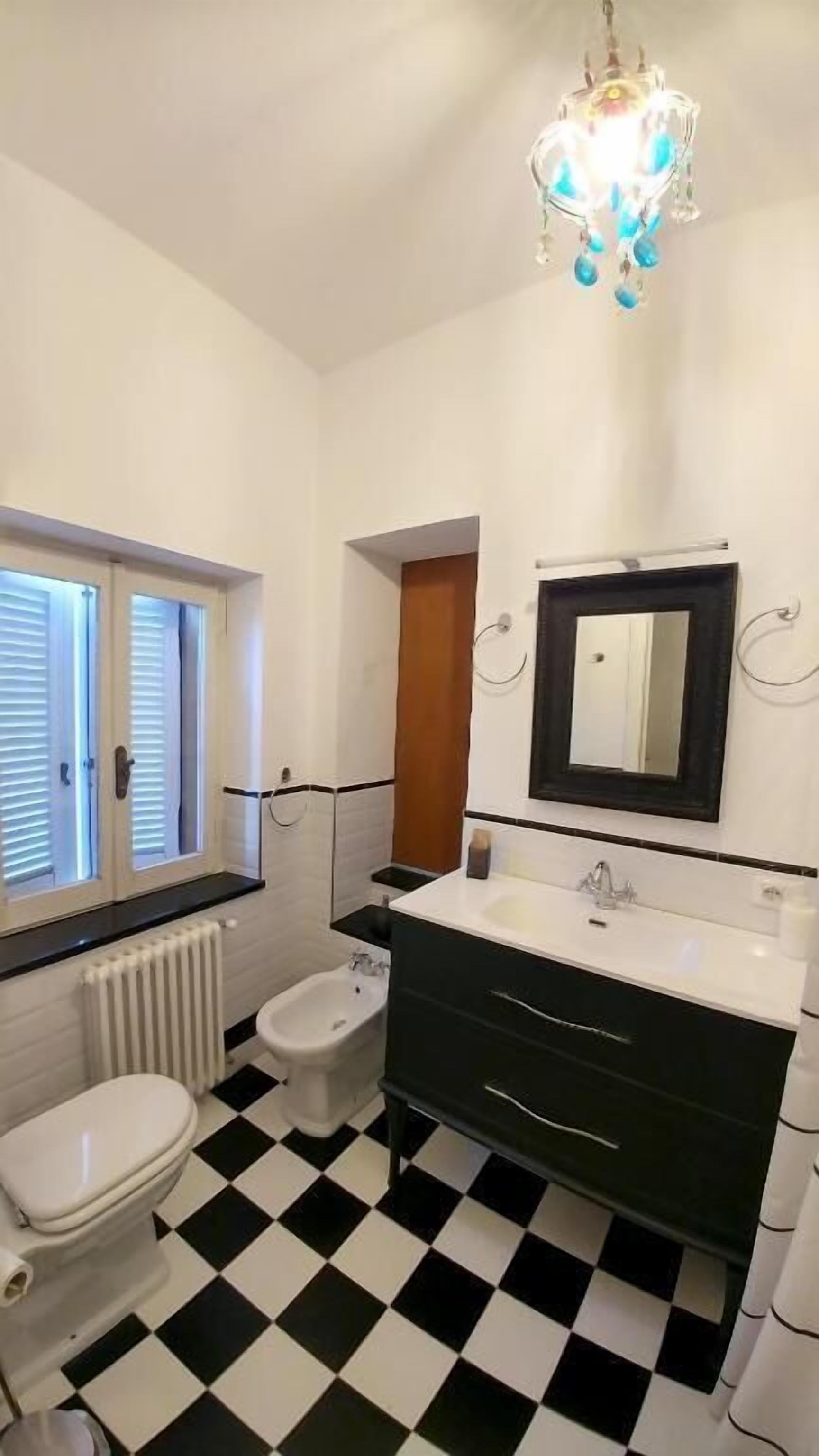 Un baño con piso a cuadros blanco y negro, un inodoro blanco, un bidé, un tocador negro con espejo y una araña.