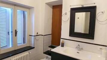 Un baño con piso a cuadros blanco y negro, un inodoro blanco, un bidé, un tocador negro con espejo y una araña.