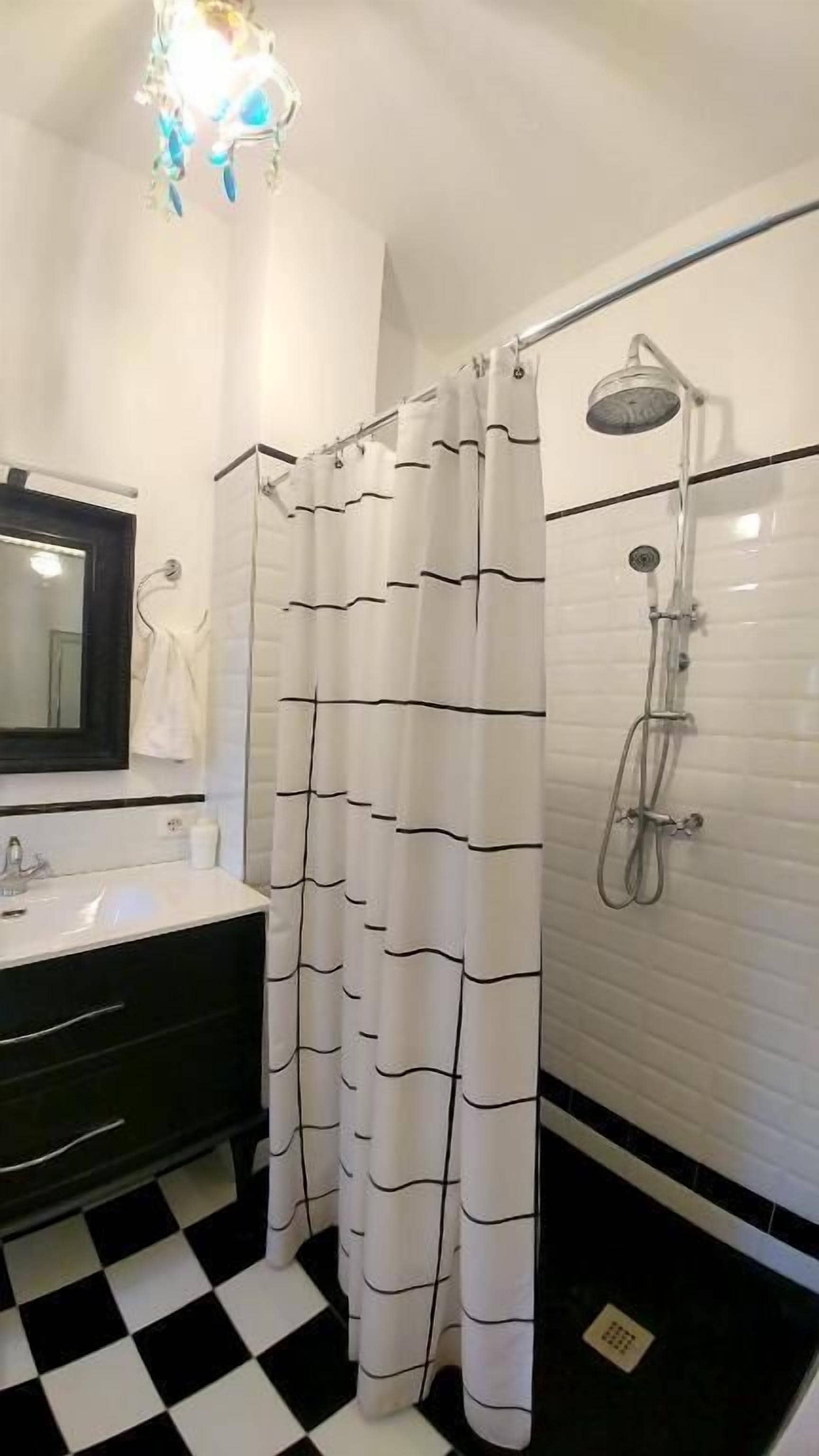 Un baño con pared de azulejos blancos, suelo a cuadros blanco y negro, ducha con cabezal de ducha manual y lavabo blanco con espejo encima.