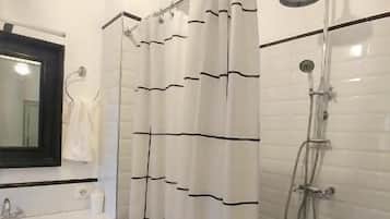 Un baño con pared de azulejos blancos, suelo a cuadros blanco y negro, ducha con cabezal de ducha manual y lavabo blanco con espejo encima.