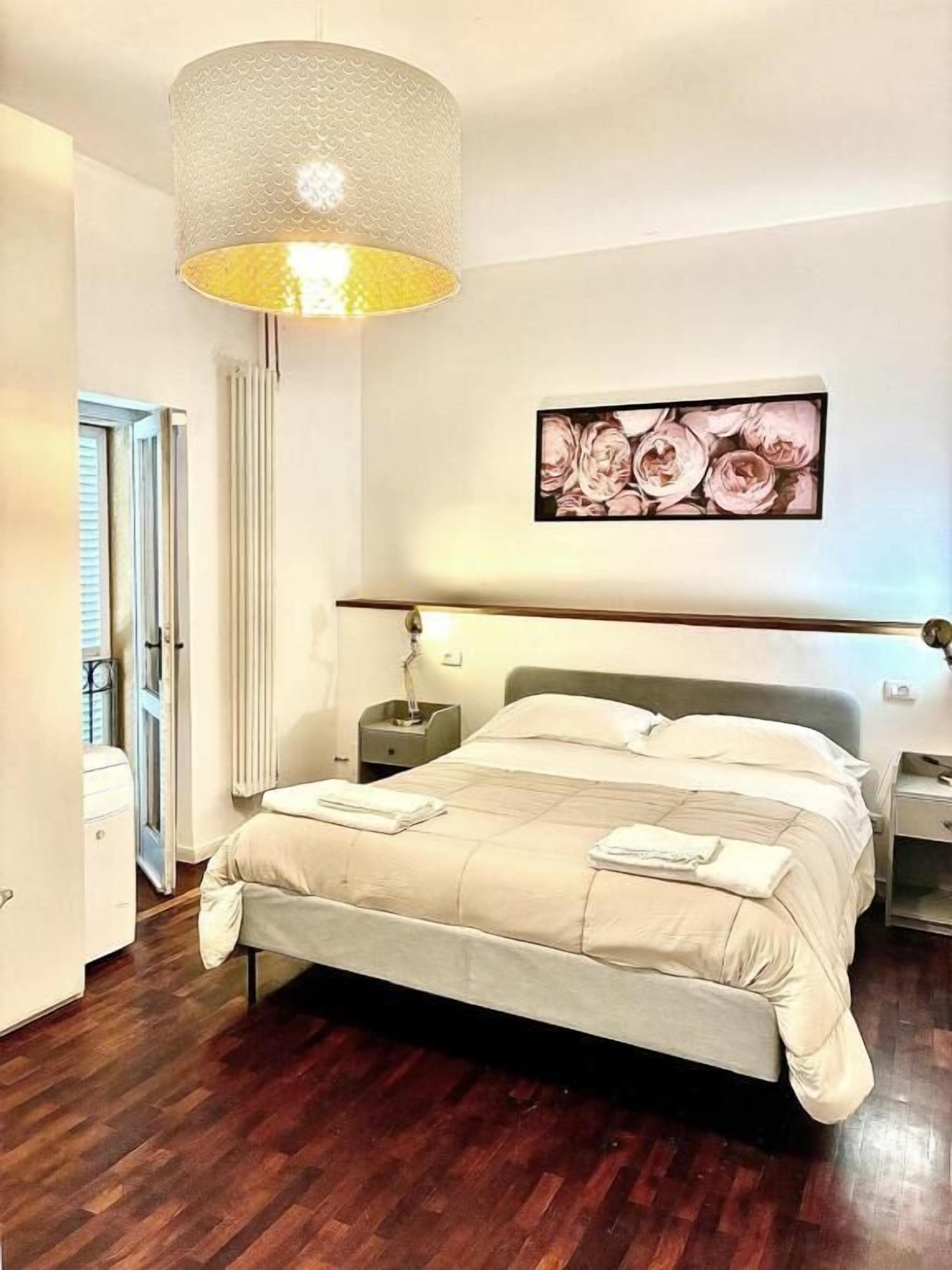 Un dormitorio con cama, mesitas de noche, lámpara, un cuadro en la pared y una puerta.