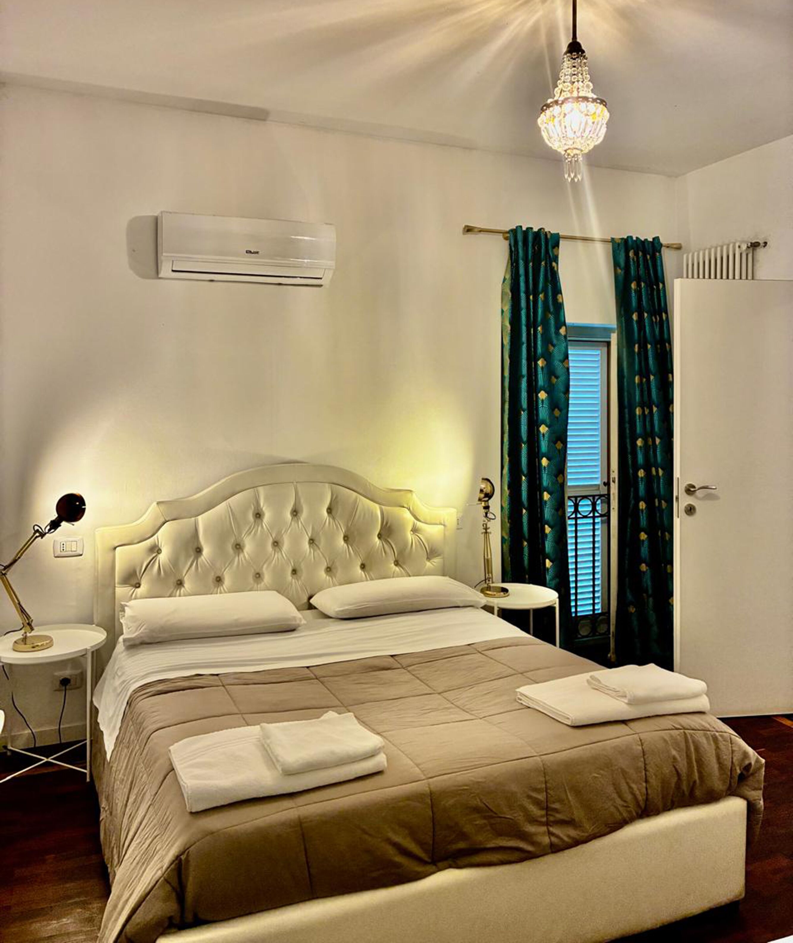 Una cama bien hecha con cabecera, lámpara de noche, equipo de aire acondicionado y una ventana con cortinas.