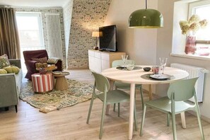 Een moderne woonkamer met een ronde eettafel, groene stoelen, een televisie op een kast en een vloerkleed met een patroon.