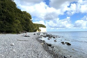 Ein Kiesstrand mit Menschen, ein bewaldeter Klippenhang und ein klarer Himmel.