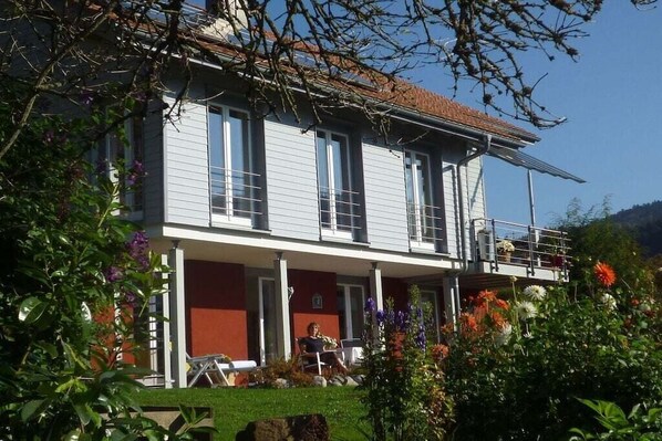 A two-story building with a balcony, a covered porch, and a garden with various flowers. - Carla Family van der Meyden (Todtmoos)