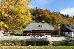 A building with a sloped roof, a stone wall in the foreground, and a forested hillside in the background. - Carla Family van der Meyden (Todtmoos)
