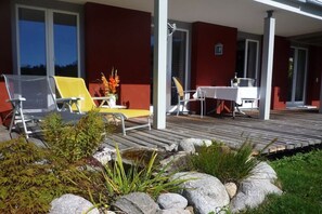 A covered outdoor area with a wooden deck, seating with a table, and a small garden with rocks and plants. - Carla Family van der Meyden (Todtmoos)
