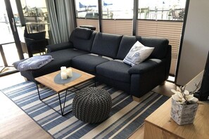 Una sala moderna con un sofá seccional gris oscuro, una mesa de centro de madera, un reposapiés redondo y una mesita auxiliar con un jarrón.