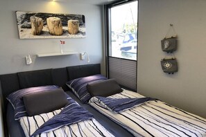 A bedroom with two beds, a window with blinds, a wall-mounted shelf, and a painting. - LaFe Flensburg Fjord (Egernsund)