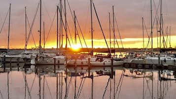 Un puerto deportivo con varios veleros amarrados, reflejándose en aguas tranquilas, y una puesta de sol al fondo.