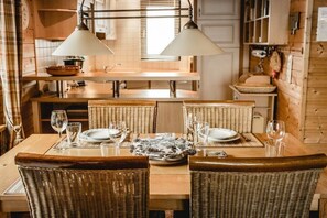 Un comedor con una mesa de madera dispuesta para cuatro personas, rodeada de sillas de mimbre, iluminación colgante y una cocina al fondo.