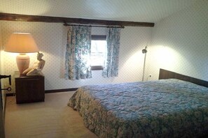 Une chambre à coucher avec un lit, une table de chevet avec une lampe, une fenêtre avec des rideaux et un mur recouvert d’un papier peint à motifs.