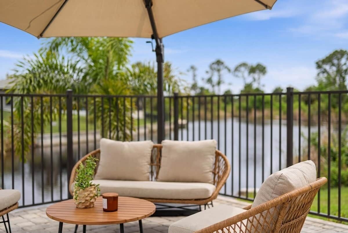 Ein Korbsessel mit Kissen, ein runder Holztisch und ein Blumentopf auf einer Terrasse mit Blick auf ein Gewässer und viel Grün.