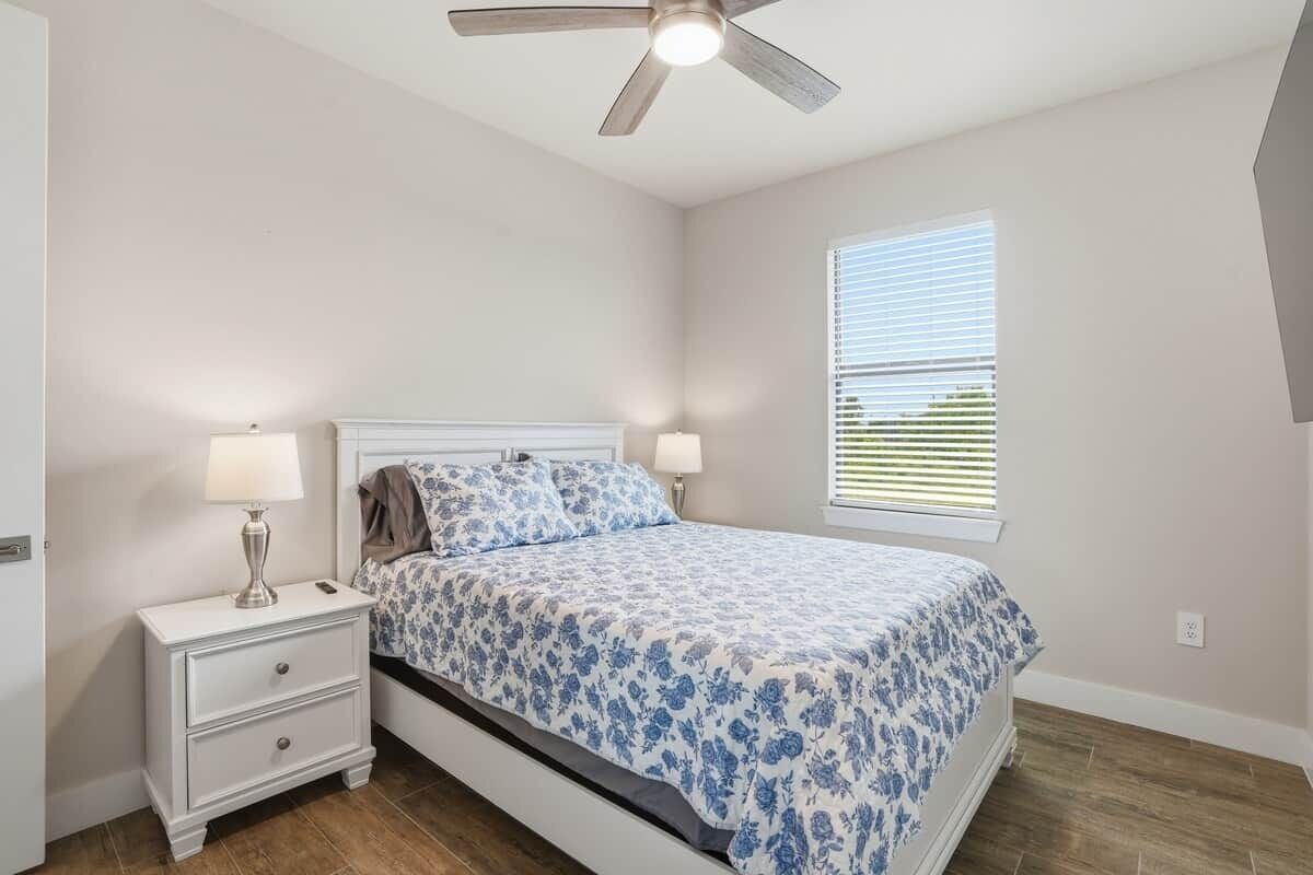 Quarto com cama, mesa de cabeceira, abajur, ventoinha no teto, janela e piso de madeira.