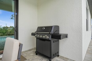 A portable grill with four burners, a side shelf, and a control panel, placed on a patio with a view of a swimming pool. - Private Accommodation in Cape Coral—Waterfront with a Pool and a Stunning View (Cape Coral)