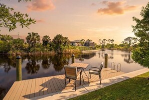 A serene lakeside setting with a wooden dock, a table, and chairs for outdoor dining, surrounded by lush greenery and a house in the background. - Private Accommodation in Cape Coral—Waterfront with a Pool and a Stunning View (Cape Coral)