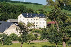 Un grand bâtiment blanc, doté de nombreuses fenêtres et d’une façade symétrique, entouré de verdure et d’arbres.