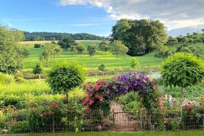 A lush garden with a variety of flowering plants and trees, a fence, and a view of a green field and trees in the distance.