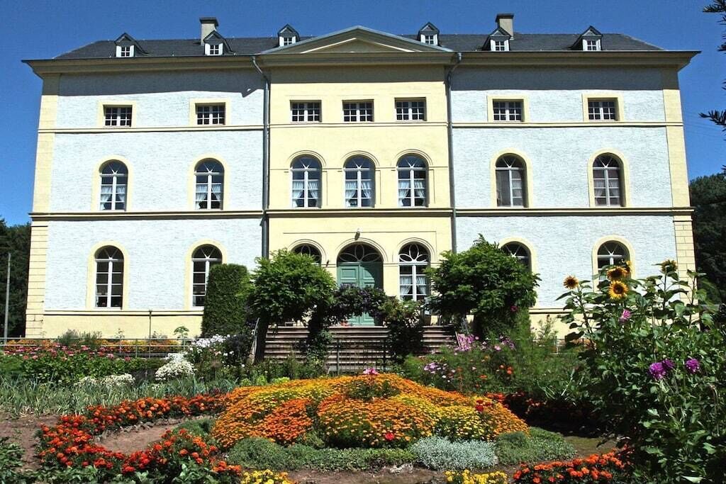 A large, multi-story building with a symmetrical facade, a central entrance with steps, and a formal garden with colorful flower beds in front.