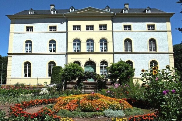A large, multi-story building with a symmetrical facade, a central entrance with steps, and a formal garden with colorful flower beds in front.