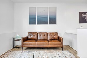Een bruine leren bank in een kamer met een witte muur, een glazen tafel met een zakdoosje en een kamerbreed tapijt met dessin.